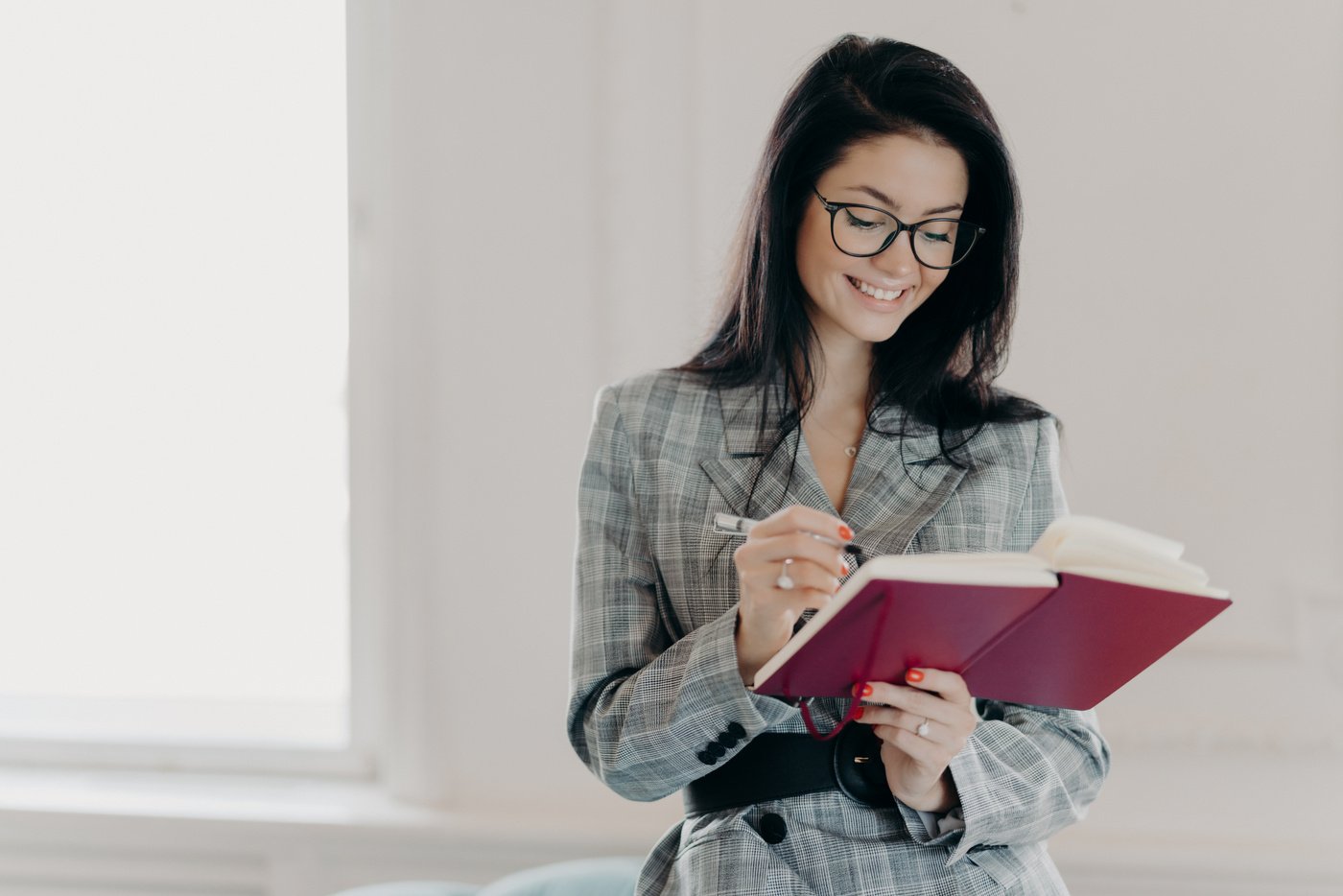 Horizontal Shot of Good Looking Woman Professor Writes down Text Notes in Notepad, Prepares for Lecture in University, Wears Transparent Glasses, Business Suit, Stands Indoor with Broad Smile